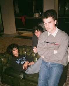 two young men sitting on top of a green couch in front of a man with his arms crossed