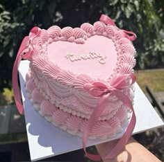 a heart shaped cake with pink frosting and ribbon on top, being held up by someone's hand