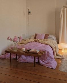 a bed with pink sheets and pillows in a bedroom next to a table with flowers on it