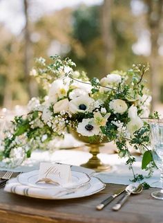 an arrangement of white flowers and greenery in a gold vase on top of a table