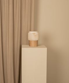 a white vase sitting on top of a wooden block in front of a beige curtain