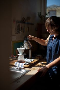 a woman pouring something into a cup on top of a wooden table in front of a window