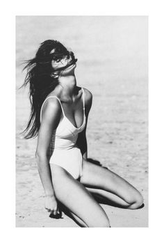 a black and white photo of a woman in a bathing suit sitting on the beach