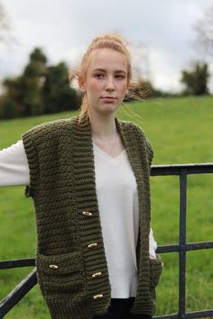 a woman standing next to a fence wearing a green knitted vest and white top