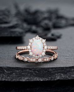 a white opal and diamond ring sitting on top of a black stone slab with flowers in the background