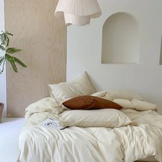 an unmade bed in a white room next to a potted plant and lamp