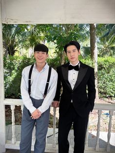 two young men in tuxedos posing for a photo on a porch with palm trees behind them