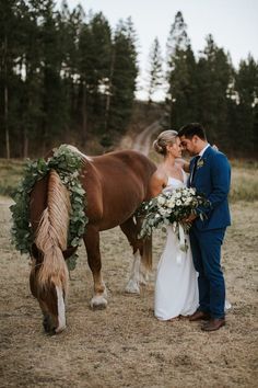 a bride and groom standing next to a brown horse with greenery on its back