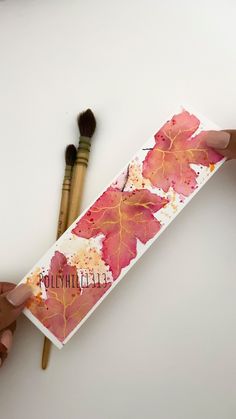 two hands are holding a card with leaves on it and paintbrushes next to it