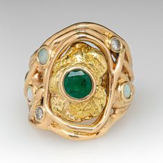 This awesome 14K yellow gold organic, freeform style ring is centered with an 18K yellow gold nugget with a bezel set, round mixed cut emerald, weighing 0.71 carats, in the middle. The shoulders are accented with a total of three (3) round, opal cabochons and two (2) round brilliant cut diamonds in bezel settings. The ring measures 23.8mm at the top, rises 4.7mm above the finger, tapering to 5.5mm wide and 22.1mm thick at the base of the shank. This ring is currently a size 7.5. The emerald shows abrasion and a light scratch/surface reaching natural inclusion. Unique Gold Emerald Ring With Bezel Setting, Unique Emerald Ring In 14k Gold, Unique Yellow Gold Emerald Ring With Bezel Setting, Unique Gold Emerald Ring Birthstone, Gold Nugget, Round Brilliant Cut Diamond, Bezel Setting, Round Brilliant, Emerald