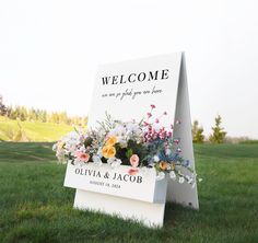 a welcome sign sitting in the grass with flowers on it's front and back