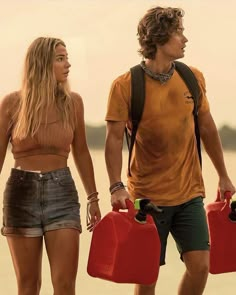 a man and woman walking on the beach with two red jugs in their hands