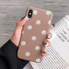 a woman is holding up her phone case with daisies on it and reading a book