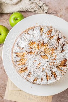 a white plate topped with a cake covered in powdered sugar next to two green apples