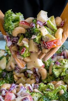 broccoli salad being held up by a wooden spoon with dressing on it and other vegetables in the background