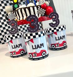 three cups with numbers and cars on them sitting on a table in front of a cake