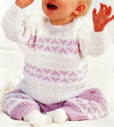 a baby sitting on a bed wearing a knitted sweater and holding her hands up in the air