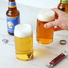 a hand holding a beer glass next to two bottles and a bottle opener on a counter