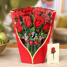 a red card with roses in a vase next to apples and a bowl of fruit