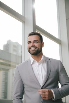 a man standing in front of a window wearing a gray suit and smiling at the camera