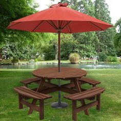 a picnic table with two benches under an umbrella in the grass near a pond and trees