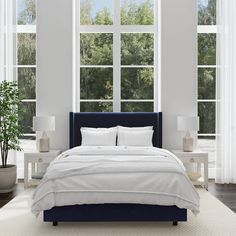 a bed with white sheets and pillows in front of two large windows that look out onto the trees