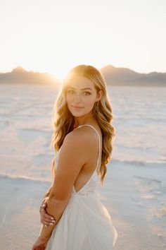 a beautiful young woman standing on top of a beach next to the ocean at sunset