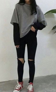 a woman wearing black ripped jeans and a grey sweatshirt with red converses stands in front of a white wall