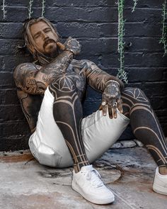 a tattooed man sitting on the ground next to a brick wall