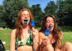 two women in bikinis sitting on the grass with blue lollipops sticking out of their mouths