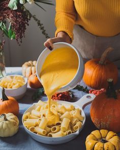 a person pouring sauce on some pasta in a white bowl next to pumpkins and gourds