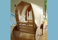 a cardboard horse drawn carriage sitting on top of a wooden floor next to a vase filled with flowers