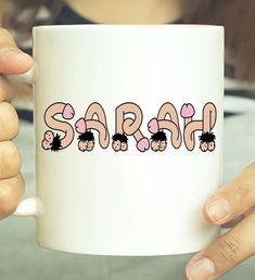 a person holding a white coffee mug with the word sarah printed in pink on it