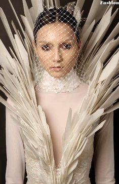 a woman with white feathers on her head and veil over her face, standing in front of a black background