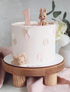 a white cake with pink frosting and two small rabbits on top, sitting on a wooden stand