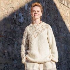 a woman standing in front of a stone wall wearing a white sweater and plaid pants