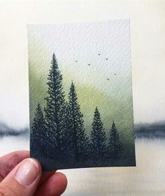 a hand holding up a piece of watercolor paper with trees painted on it and birds flying in the sky