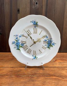 a white clock with blue flowers on it sitting on top of a wooden table next to a wall