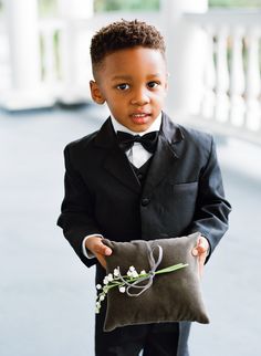 a young boy in a tuxedo holding a pillow with a flower on it