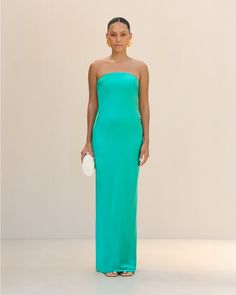 a woman in a strapless green dress stands against a white wall and holds a white purse