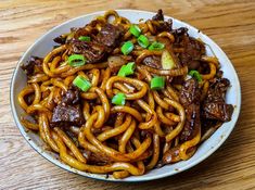 a white bowl filled with beef and noodles on top of a wooden table next to chopsticks