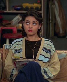 a woman sitting on top of a couch holding a magazine in her hand and looking at the camera