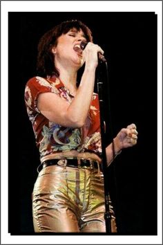 a woman singing into a microphone while wearing metallic pants and a floral shirt on stage