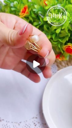 a woman is holding a ring in her hand with a pearl on the end and a flower pot behind her