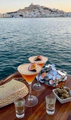 two martinis with olives and other food on a table next to the water