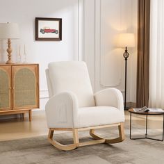 a white rocking chair sitting in a living room next to a table with a lamp on it