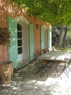 an outdoor table and chairs under a tree with green shutters on the outside wall