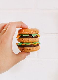 a hand holding a hamburger with green and black cheese on it, in front of a white brick wall