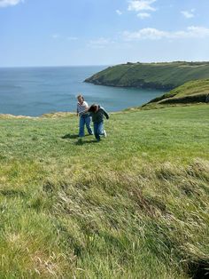 two people standing on top of a lush green hillside next to the ocean and grass