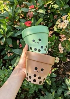 two cups with faces painted on them are held up in front of some bushes and flowers